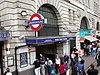 A street filled with people in front of a light grey building that has variously coloured signs protruding from it stating a variety of different things