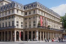 Farbige Eckansicht von einem viergeschossigen Gebäude mit Arkaden im Erdgeschoss. Die Fenster haben Sprossen und Steingitter. Am rechten ersten Geschoss ist ein langer Eisenbalkon mit zwei Sträuchern und drei horizontalen Bannern.