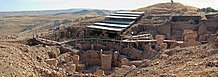 Roughly built stone walls surrounding T-shaped stone pillars under a modern steel walkway and roof in a hilly landscape