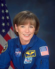 Formal portrait of astronaut in a flight suit displaying mission patches with an American flag in the background