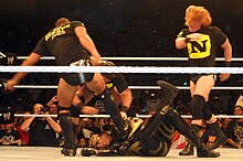 Sur un ring, trois membres du Nexus — Justin Gabriel (de dos), Wade Barrett (masqué) et Heath Slater — attaquent le catcheur Goldust, à terre.