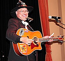 A man wearing black clothing and a black hat, holding a guitar and standing behind a microphone