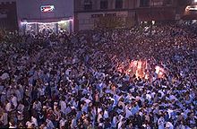 A large gathering of people on a street with a bonfire in the right side of the image.