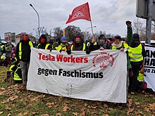 At an outdoor demonstration in Riesa, Germany, 6 Tesla workers are carrying a banner that says "Tesla Workers gegen Faschismus" (Tesla workers against fascism). There is an IG Metall flag in background, and also on the banner itself. The faces of people holding the banner are blurred.