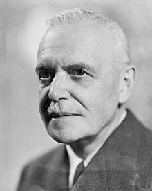 Portrait en noir et blanc d'un homme âgé avec des cheveux blancs, une moustache fine et vêtu d'un costume. L'image est cadrée sur son visage, qui pose en regardant légèrement vers la gauche.