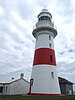 Low Head Lighthouse, a white-painted lighthouse with a red band halfway up and a weathervane on top, a white-painted single storey building with chimneys to the left and a grey-painted generator shed to the right