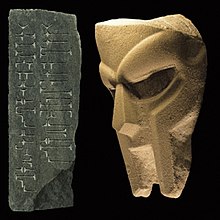 A stone or concrete rendition of the modern MF DOOM mask, facing a stone with abstract symbols etched into it.