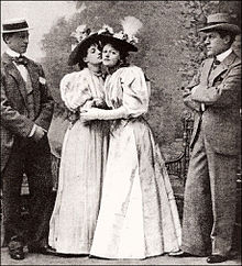 group of four young white adults, standing in an outdoor stage setting, two men and two women, in late Victorian dress, all wearing hats. The two women are in the middle and are cautiously embracing each other, while the men look on.