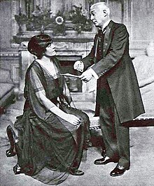 Dark haired white woman, seated in a drawing room with older, white haired white man standing before her and showing her a piece of paper