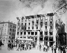 A black and white photograph of a burning building, with a small crowd watching.