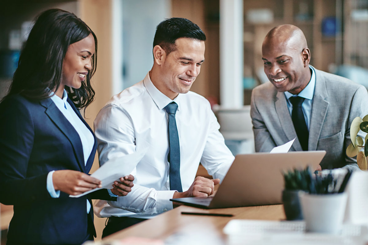 Three smiling people in business attire, standing at a table, all looking at the same laptop