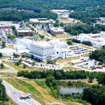 An aerial photograph of the NASA Goddard Visitor Center, with white and brick rectangular buildings peeking out from among dense green trees behind a tall white rocket. The rocket stands in the center of a tan paved circle and is shaped like a pencil with a long, slim tower and a slight bulge at the top. Several smaller red, white, and yellow rockets are displayed on their sides around the paved circle, with another rocket standing in a smaller, white paved circle near the trees.