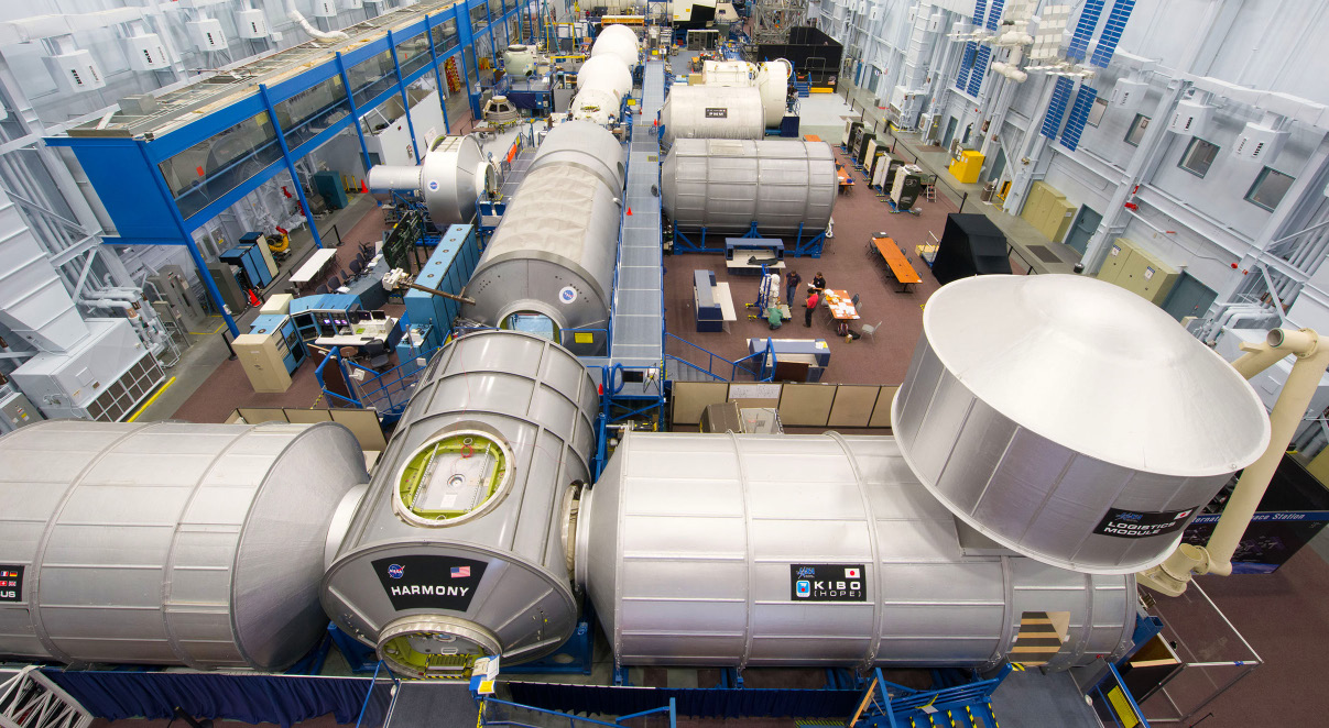 Silver cylinders that serve as mockups of the International Space Station are conjoined in a T shape on the floor of a large building