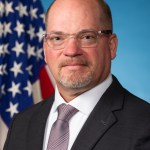 Chad Brown in glasses wearing a suit and tie poses for a headshot photograph in front of an American flag and blue backdrop.