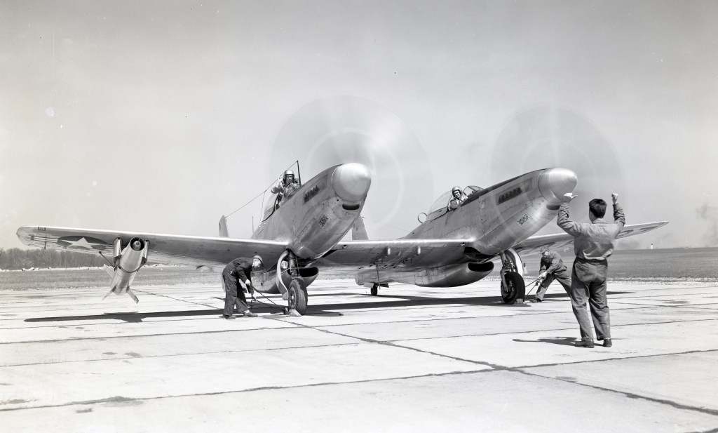 Aircraft with two pilots inside, propellers in motion, watch as ground crew guide them on taxiway.