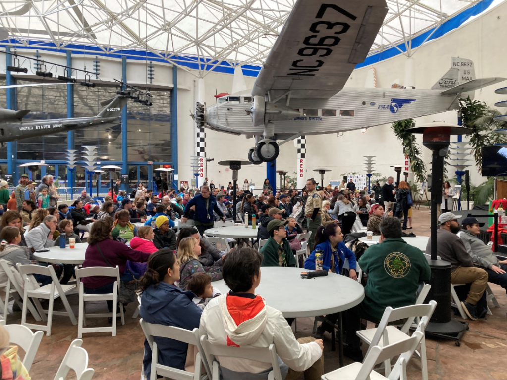 A large gathering of Scouts sitting around tables, looking off the right side of the picture toward a stage.