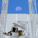 A close-up photo of a Deep Space Network antenna. Behind the antenna, the Moon can be seen clearly on a bright blue sky.