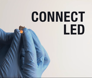 Gloved hands hold an LED light lit by battery made from pennies and the words "Connect LED"