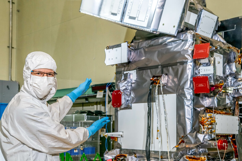 A man stands to the left of the image, seen from the waist up. He is wearing glasses, blue latex gloves, a white mask covering his nose and mouth, and a white clean suit that covers his arms, torso, and head. He gestures to the right of him, holding both hands out towards a large box-shaped item - the PACE spacecraft. The spacecraft is covered in wires, other mechanical sensors and items, and a silver-colored foil-like material.