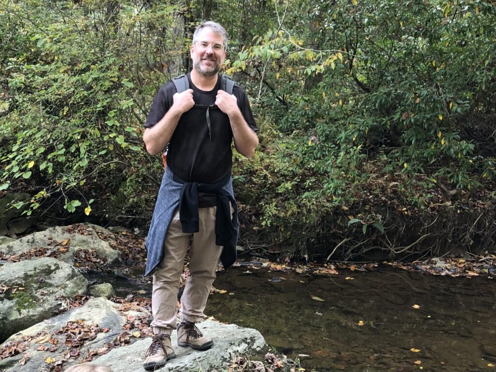 A man stands in the left of the image wearing light brown pants and a black T-shirt. He stands on a rock next to a small body of water. In the background there are green leaves and foliage. The man stands looking at the camera.