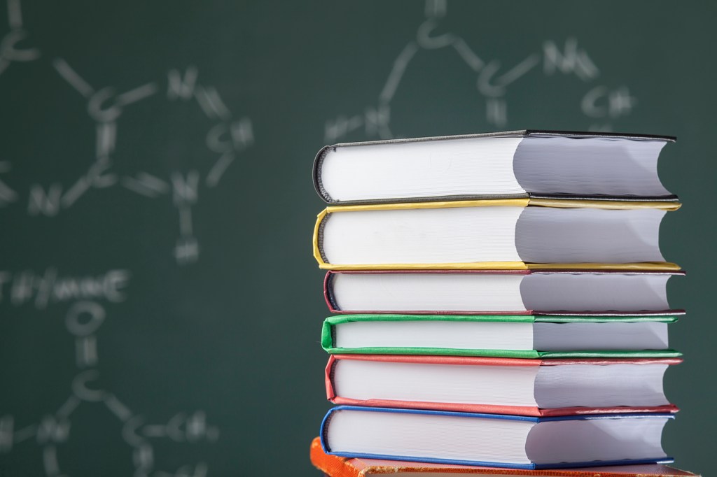 Stack of multicolored books on blackboard with organic molecules written on.The books are placed on the right side of frame.Human DNA formulas are written on board.The focus is on the books and blackboard is blurred.The photo was shot with a full frame DSLR camera in studio lighting.