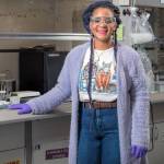 Young woman with safety glasses, dressed in t-shirt, jeans, and purple fuzzy sweater with purple gloves, faces camera while standing in laboratory