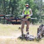 A drone flies in the foreground while a person wearing a hat, sunglasses, and yellow safety vest pilots the drone in the background, with a forest behind.