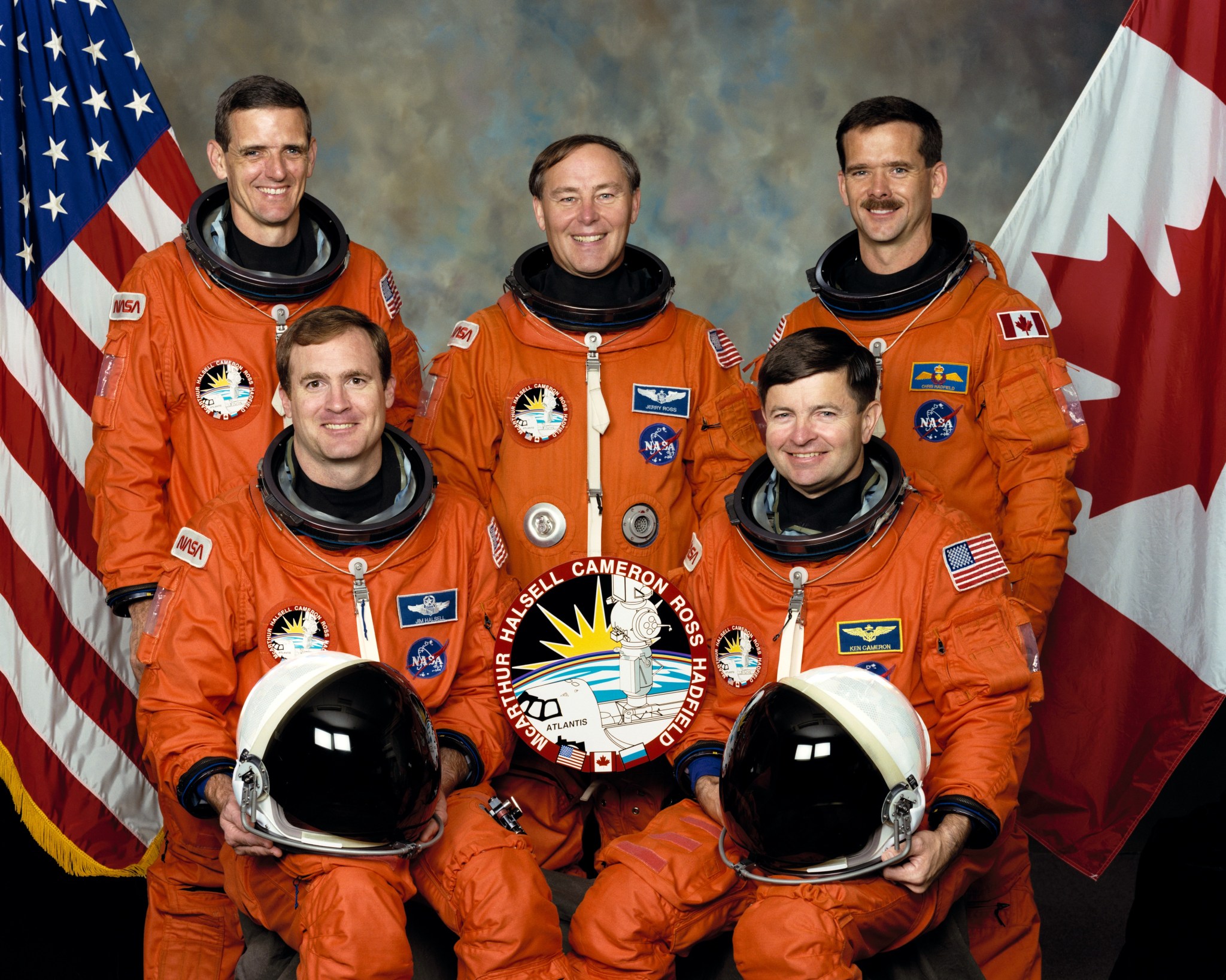 STS-74 crew members wearing their orange launch and entry suits with both the American and Canadian flags in the background.