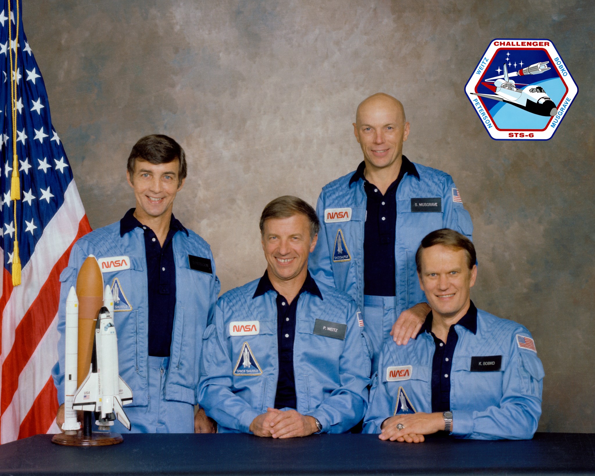 Four men in blue NASA outfits pose with model shuttle and U.S. flag