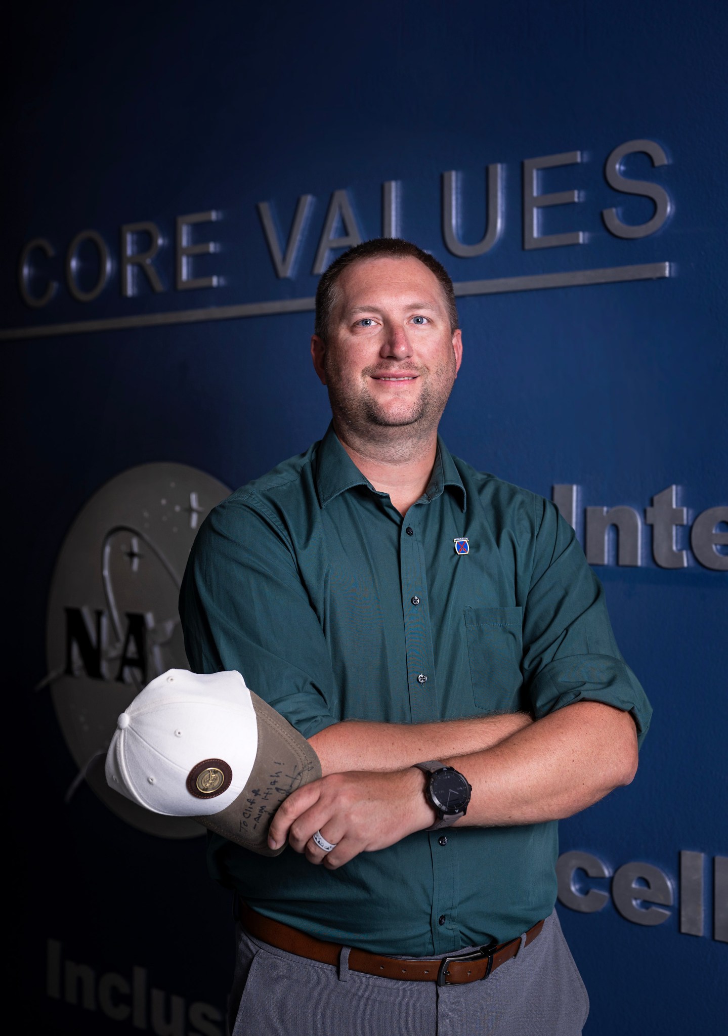 Luke Bingaman, a white man, stands with his arms crossed with a military hat in his hand.