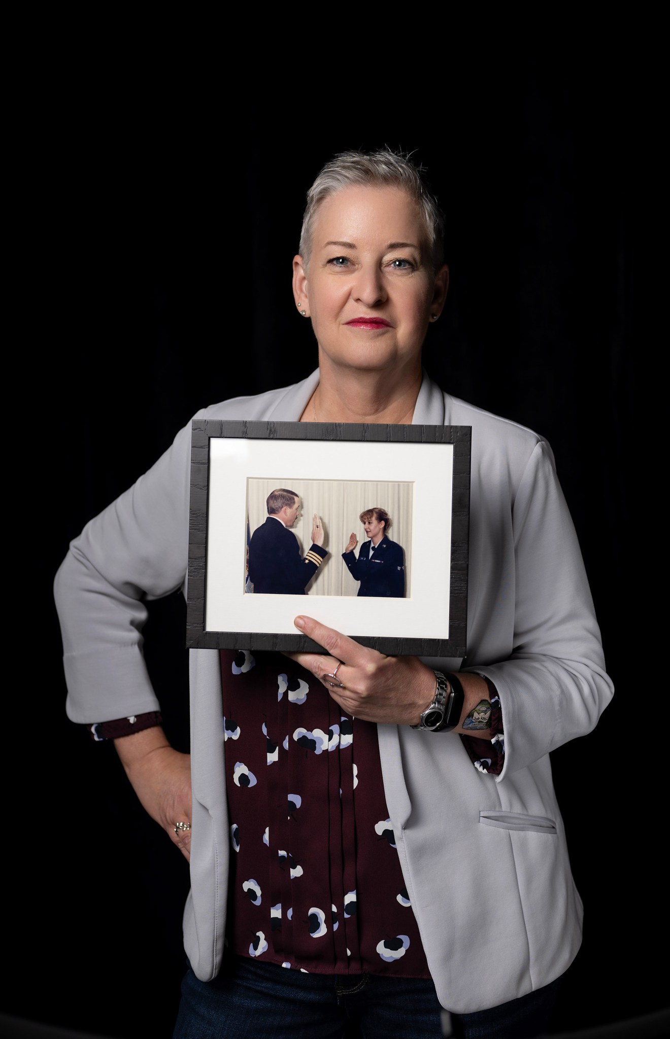 A woman with short hair stands holding a photo of herself in the military.