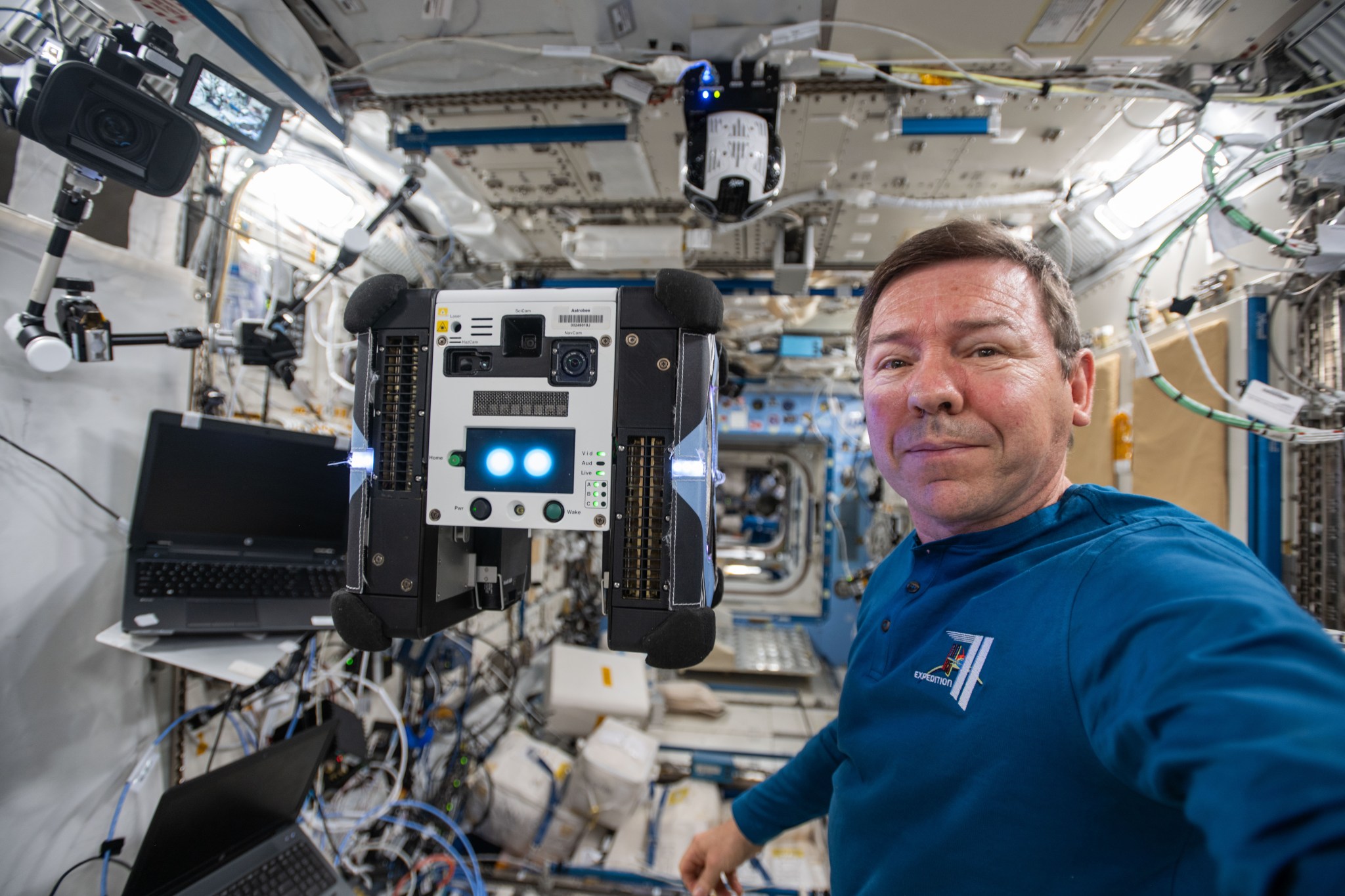 Barratt, wearing a long-sleeved blue shirt with an expedition patch on its chest, faces the camera. Just to his right floats Bumble, a cube-shaped robot about the size of a toaster oven. Its sides are black with a white panel in the middle that has camera lenses and two bright blue lights near the bottom. There is a laptop to his right.