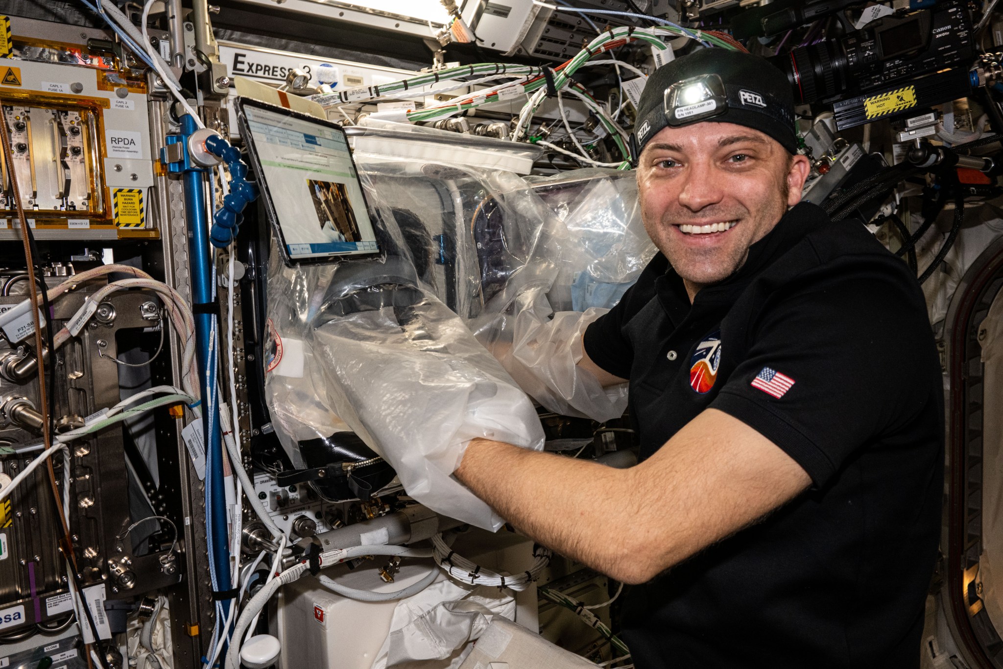 Dominick, wearing a headlamp and black polo shirt with a US flag decal on the left sleeve, smiles at the camera. His arms are inside the plastic sleeves of a portable glovebox, and a laptop is visible just above and to the left of the glovebox.