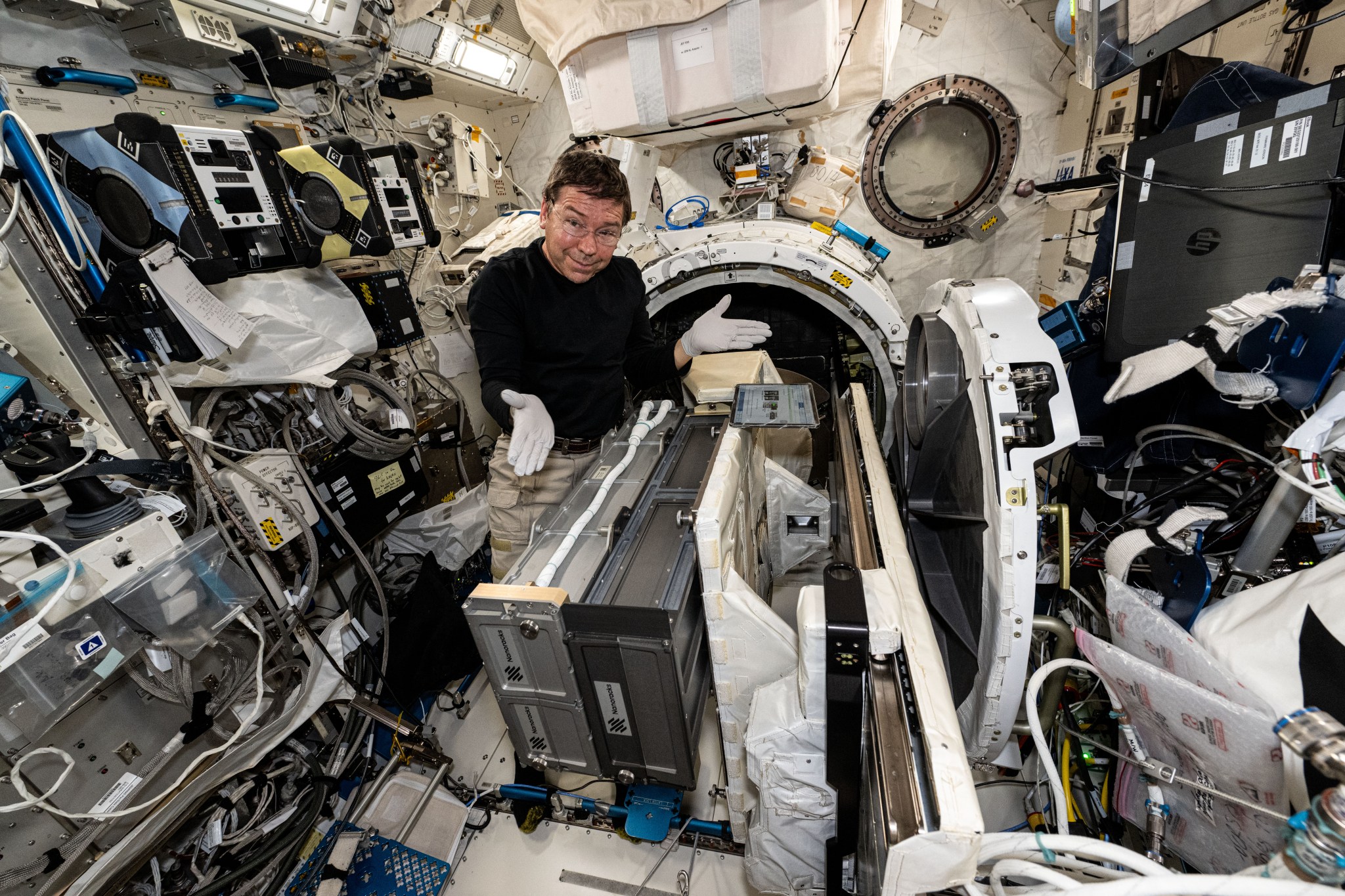 Barratt, wearing a long-sleeved black shirt, khaki pants, and white gloves, looks at the camera and gestures at the hardware in front of him, two large rectangular drawers mounted sideways on a rack extending out from a circular hatch behind him. The open door of the hatch is to his left, and the blue and yellow boxy Astrobee robots are attached to their docks on the wall to his right.