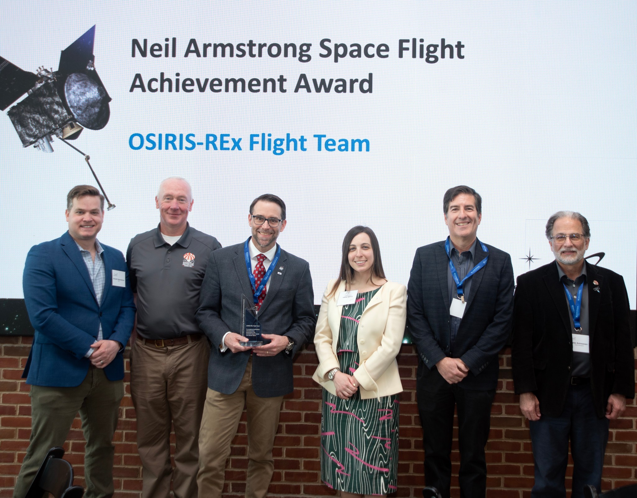 Six members of the OSIRIS-REx team pose for a photo in front of a screen that says: "