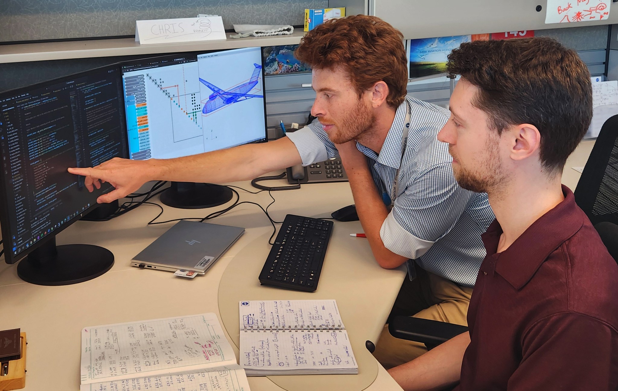 Two men work at a desk in a NASA office as one points to some Aviary computer code displayed on a monitor. A picture of a future aircraft design appears on a neighboring monitor.