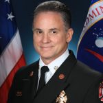 A man in a suit jacket and tie is centered in front of a background of two flags.