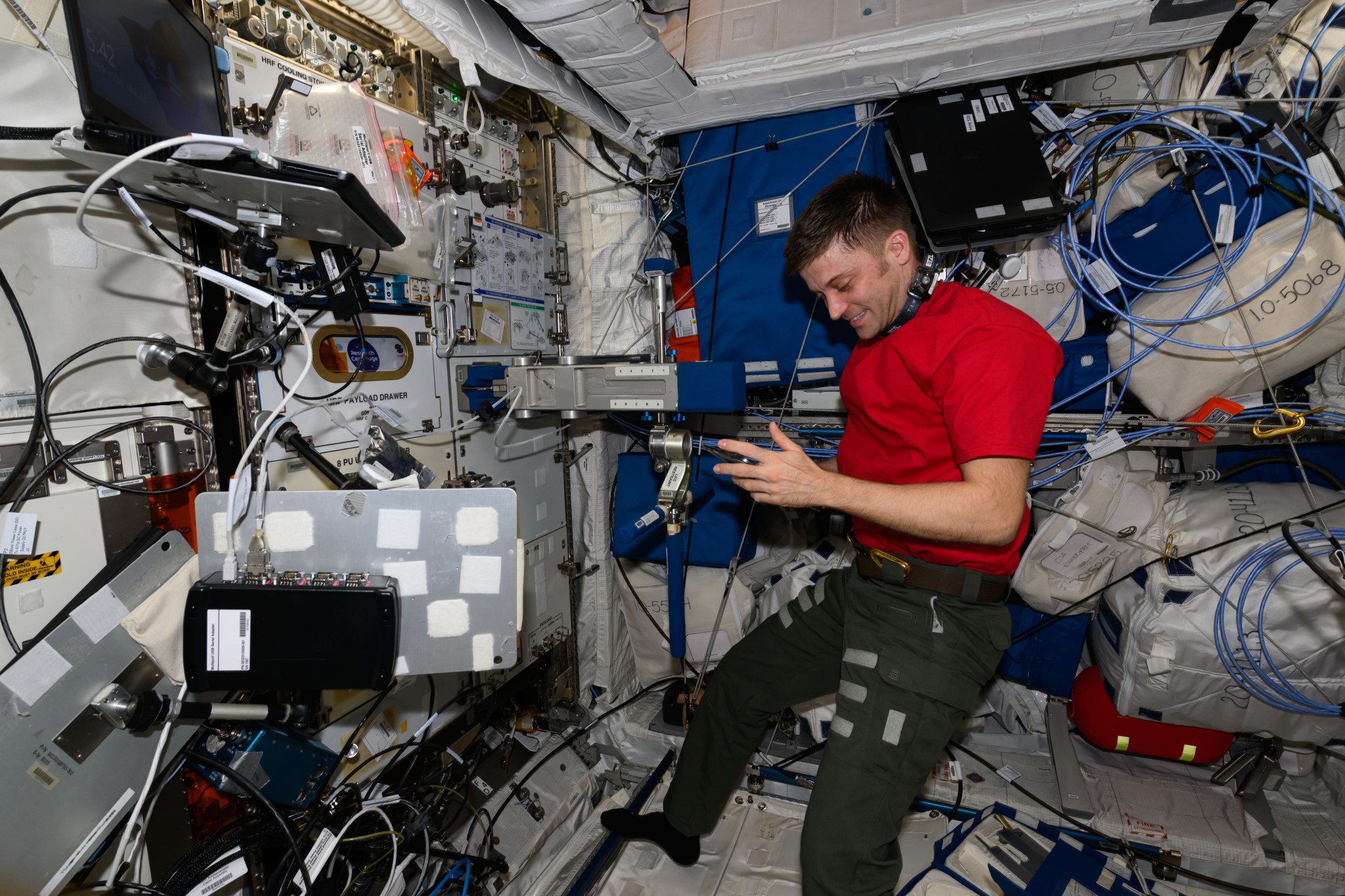 Dominick, wearing a red t-shirt and dark green pants, is looking down and smiling. In front of him is a square silver brace with blue padding on its end with silver arms extending above and below it, also with blue padding. Above the brace is a large schematic poster showing how to use the device, and a laptop is mounted off to the left.