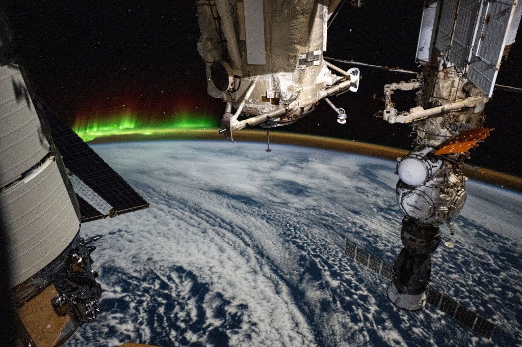 An aurora and an atmospheric glow crown Earth's horizon in this photograph from the International Space Station as it orbited 266 miles above a cloudy Indian Ocean southwest of Australia. In the foreground, from left, are a portion of Northrop Grumman's Cygnus space freighter, the Rassvet module, and the Soyuz MS-25 crew ship docked to the Prichal docking module which is itself mated to the Nauka science module.