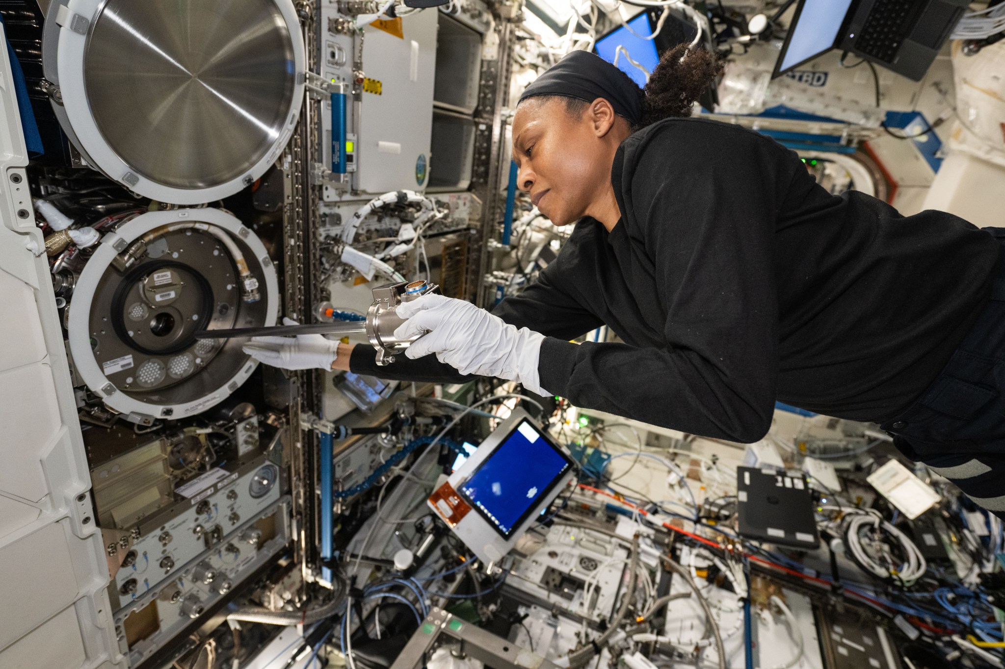 Wearing a long-sleeved black shirt and white gloves, Epps is smiling and holding a silver soup-can-sized canister in her left hand and guiding a long silver rod attached to it into an opening in the Material Science Laboratory, a metal circular device with white plastic edging.