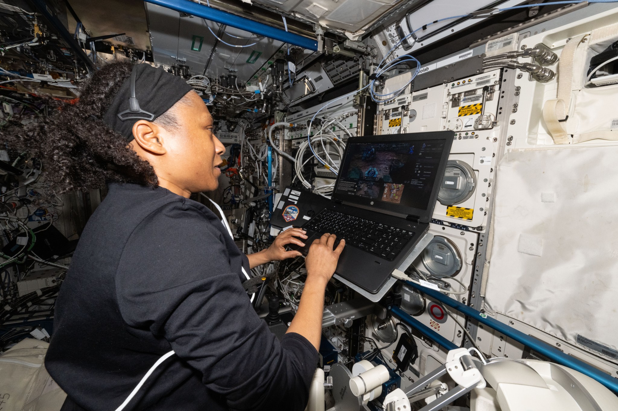 Epps wears a long-sleeved black shirt and a headset and uses her right hand to interact with a laptop mounted to the wall in front of her. On the screen is a simulation of a robot operating on a planetary surface.