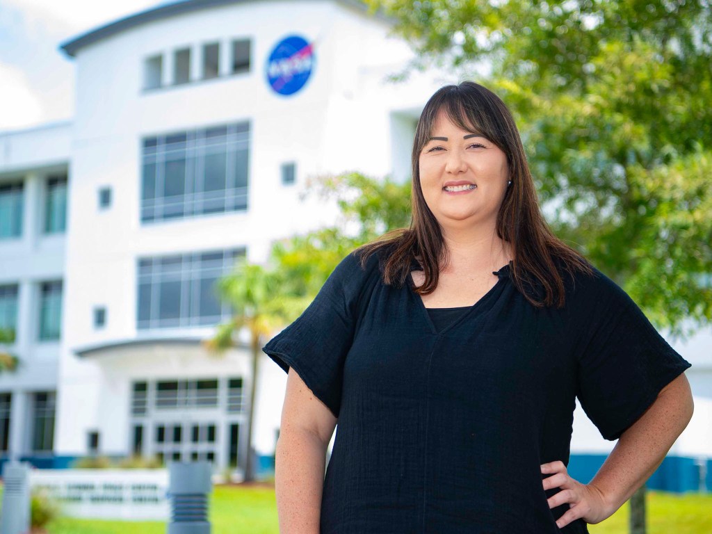 Kim Johnson, wearing a black blouse, poses near a tree outside of Building 1111 at Stennis Space Center which can be seen in the background.