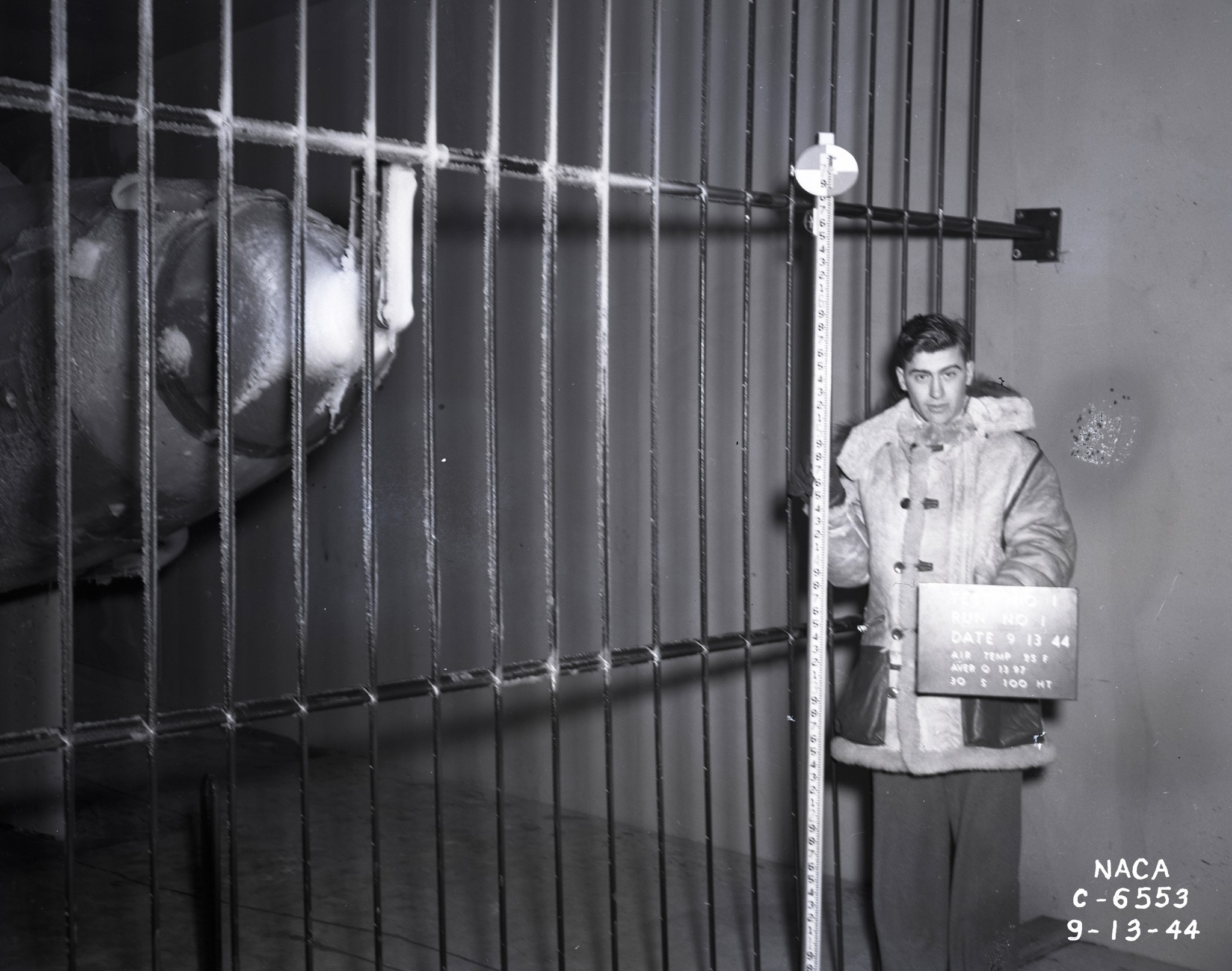 A black-and-white photo of a man who stands inside the Icing Research Tunnel beside spray bars and the nose of a plane that is covered in ice. He is wearing a large fur-lined coat and holds a tall measurement stick and a sign that says, “Test No 1, Run No 1, Date 9-13-44” and provides other test information.