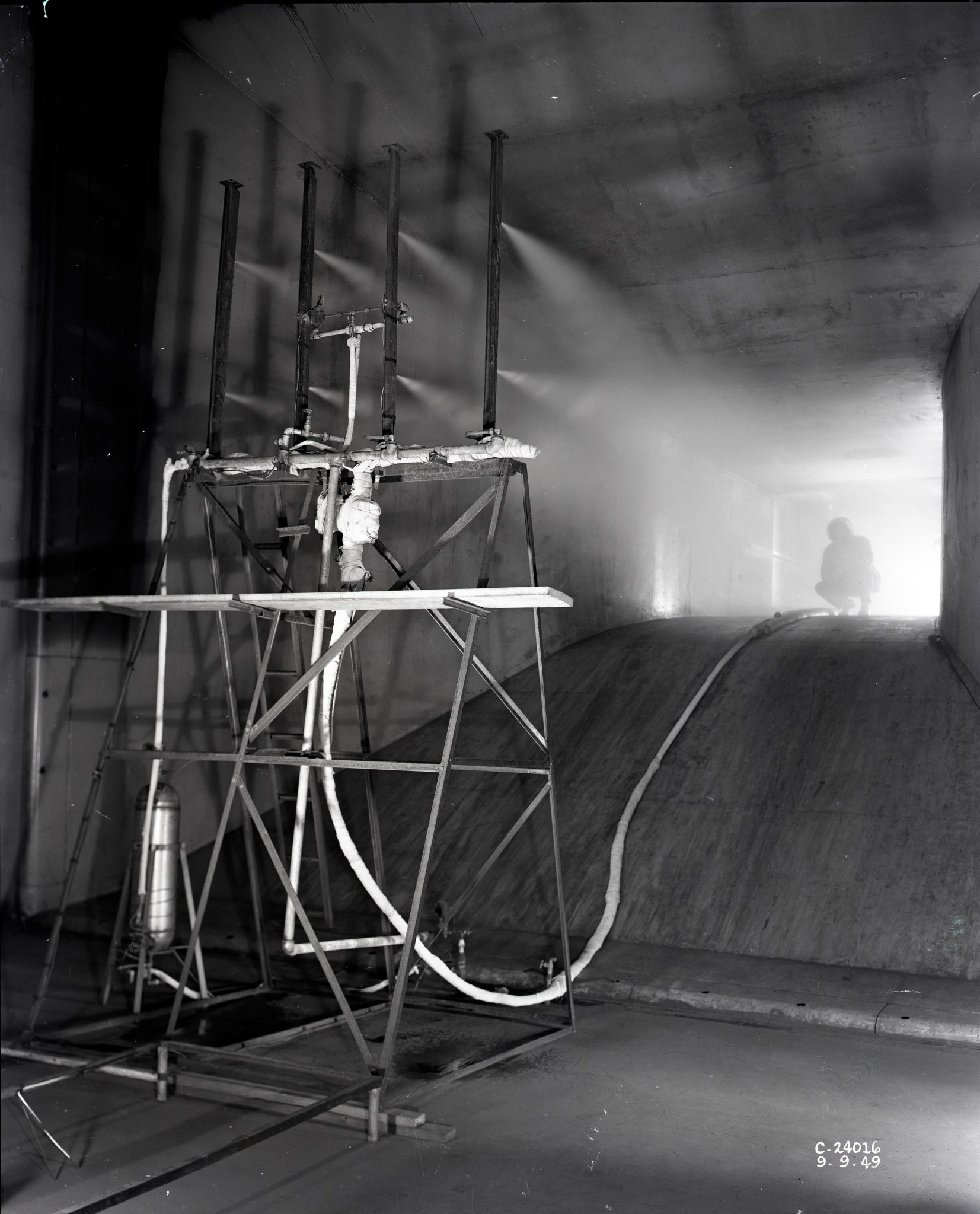 A black-and-white photo of the Icing Research Tunnel’s water spray system. Water sprays from equipment that includes multiple bars and tubes. A person crouches inside the tunnel in front of the stream of droplets.