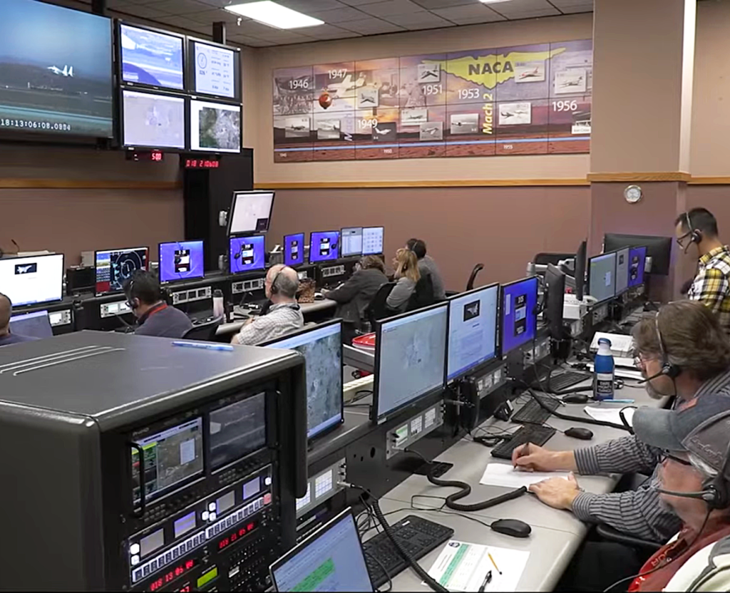 A group of people sitting in two rows in front of computer screens while watching another set of monitors on the wall in front of them.