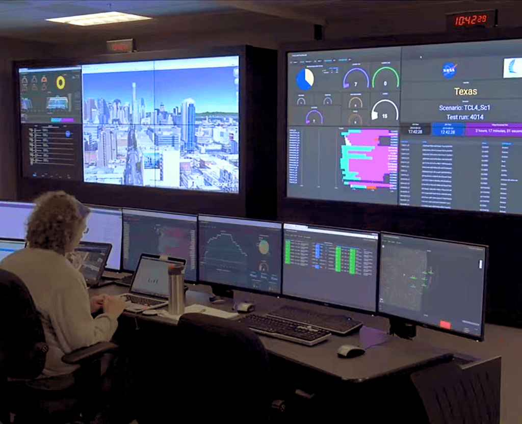 A person works at a laptop with several large monitor on the wall at front.