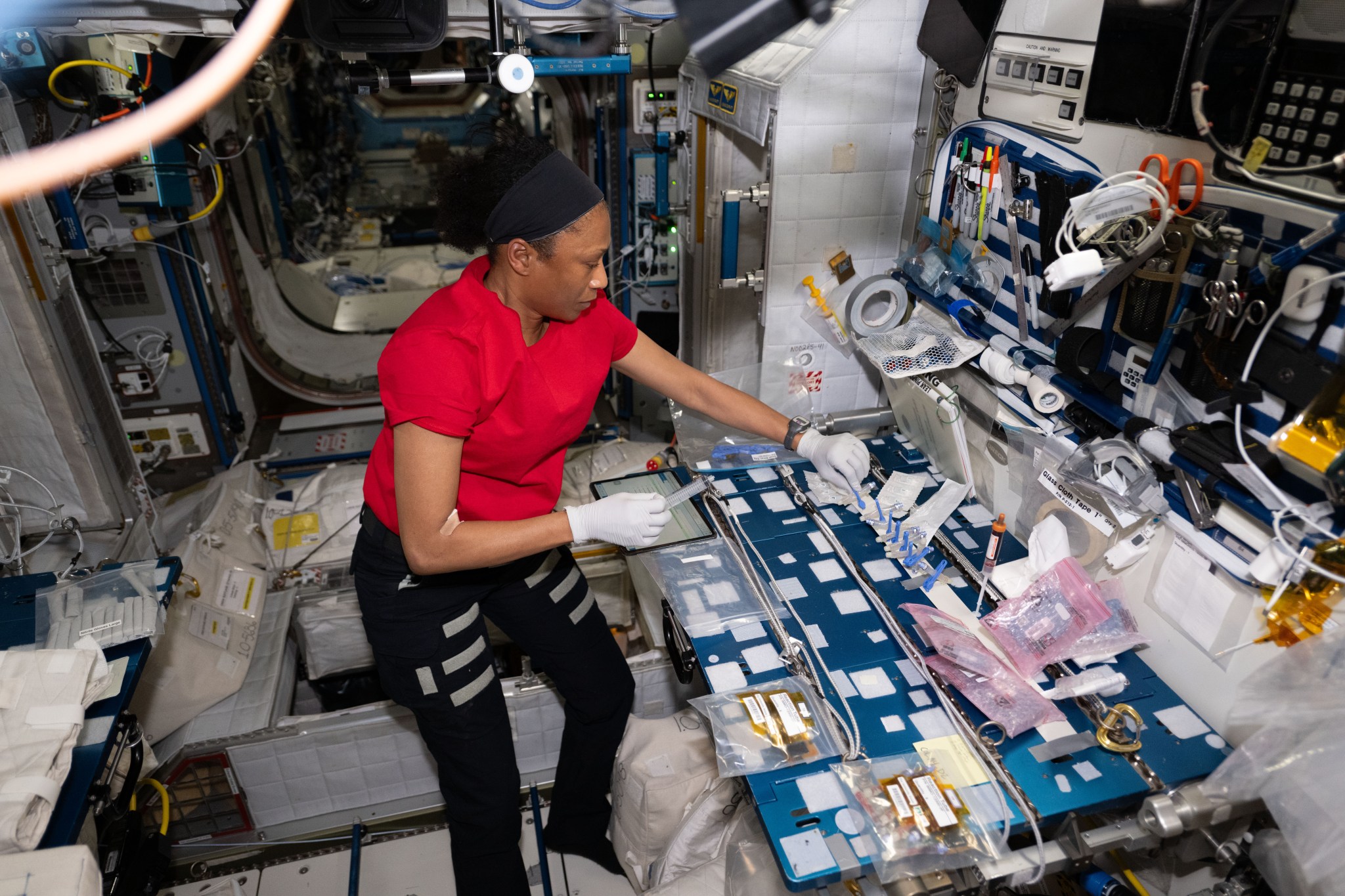 Epps wears a red t-shirt, black pants, and white gloves. She is holding a large syringe in her right hand and is using a blue clip in her left hand to attach a sample tube to the blue lab work table. There are multiple plastic bags across the front of the table and a cluster of pink bags on its upper right. The wall behind it holds various supplies, such as tape, scissors, and pens.