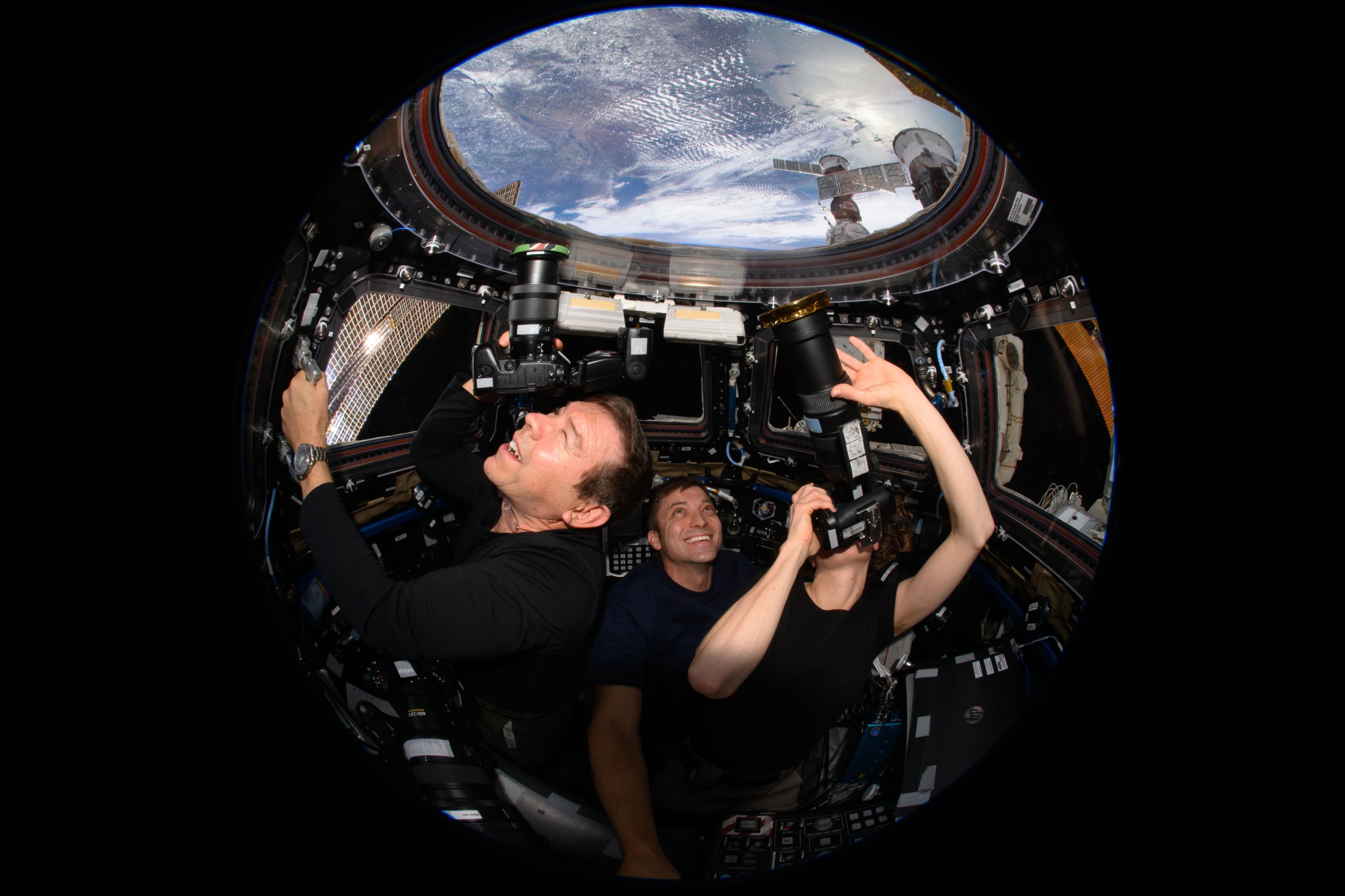 Three astronauts, all wearing black, are facing the camera in the cupola, where Earth is visible through the window above them. Barratt, on the left, holds a handrail with his left hand and points a camera toward Earth with his right hand, squinting his left eye. Dominick, center, smiles up at the window, and O’Hara, on the right, uses both hands to point a camera with a large lens at Earth. The camera obscures her face.