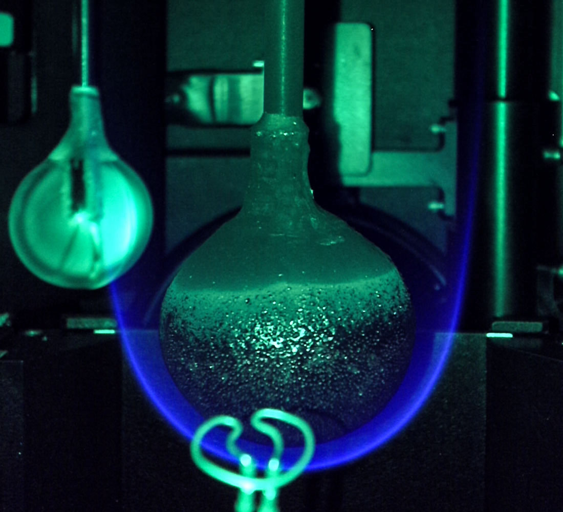 An acrylic sphere hangs from a rod in the middle of this image, a bright blue flame arcing around the bottom of it. The sphere is mottled black on its lower two-thirds and smooth near its top. A greenish, unburned sphere hangs off to its left, and a bright green wire coil (the igniter) is visible in the lower foreground. The entire image is tinged green.