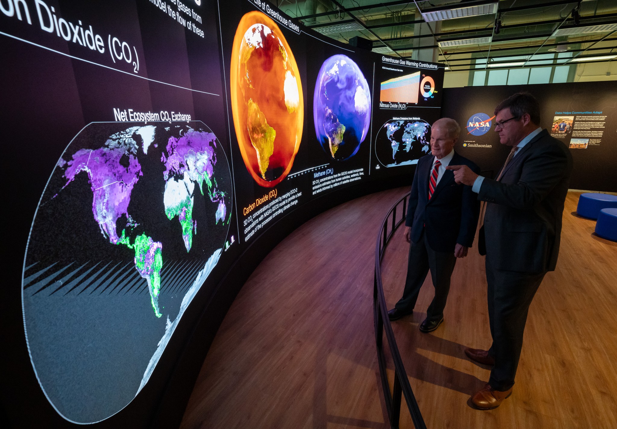 Two men in suits stand behind a short curved barrier. They look at a large curved screen that spans the room. The screen shows information, maps, and other graphics that relay information about carbon dioxide. The man on the right is pointing at the display.